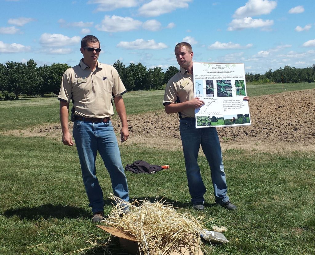 Two men presenting poster next to agricultural field.