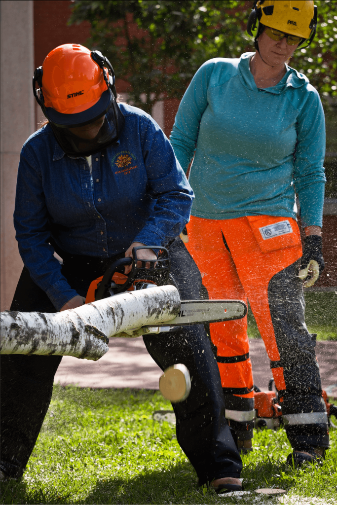 Woman in helmet and chaps uses chainsaw to cut cross-section of tree limb, instructor stands behind her