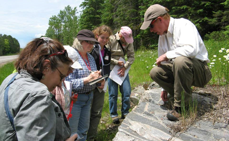 Man stands in front of group of people analyzing a rock in the outdoors.