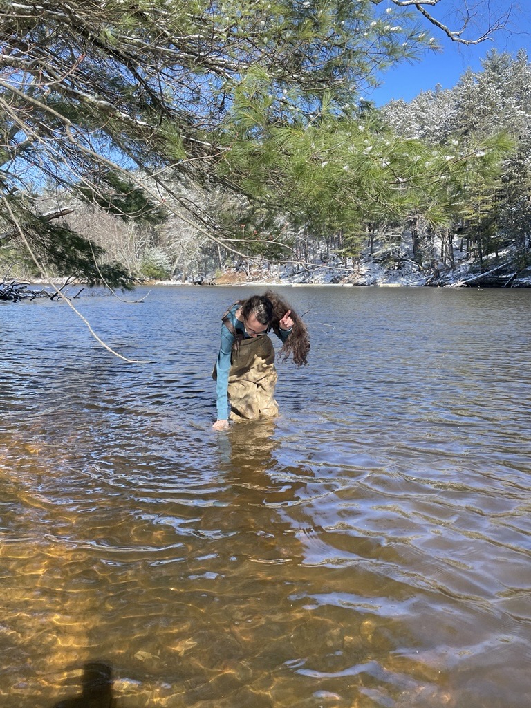 A student in waders standing in the Wisconsin River collecting a water sample