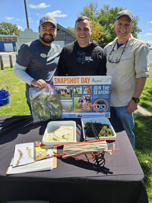 Three men standing at a table with a Snapshot Wisconsin sign, and three types of aquatic invasives species - plants and mollusks in front of them.