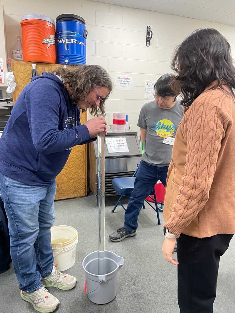 Two people watching a third do a water experiment with a tall clear tube of water in a bucket