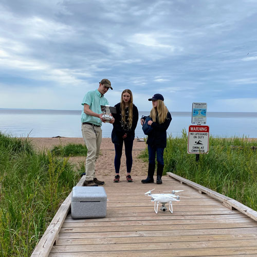 A man teaching two girls how to use a drone along a lake shoreline