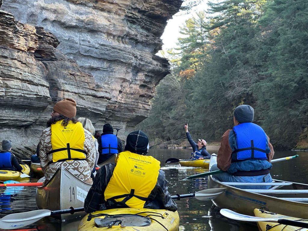 An instructor in a kayak pointing to an outcrop. A group of people in kayaks and canoes listen.