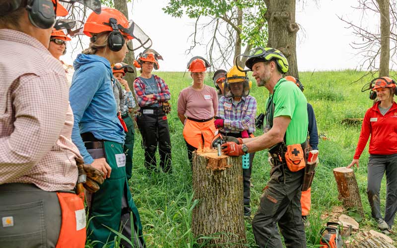 Several women dressed in chainsaw safety gear learning how to use a chainsaw safely