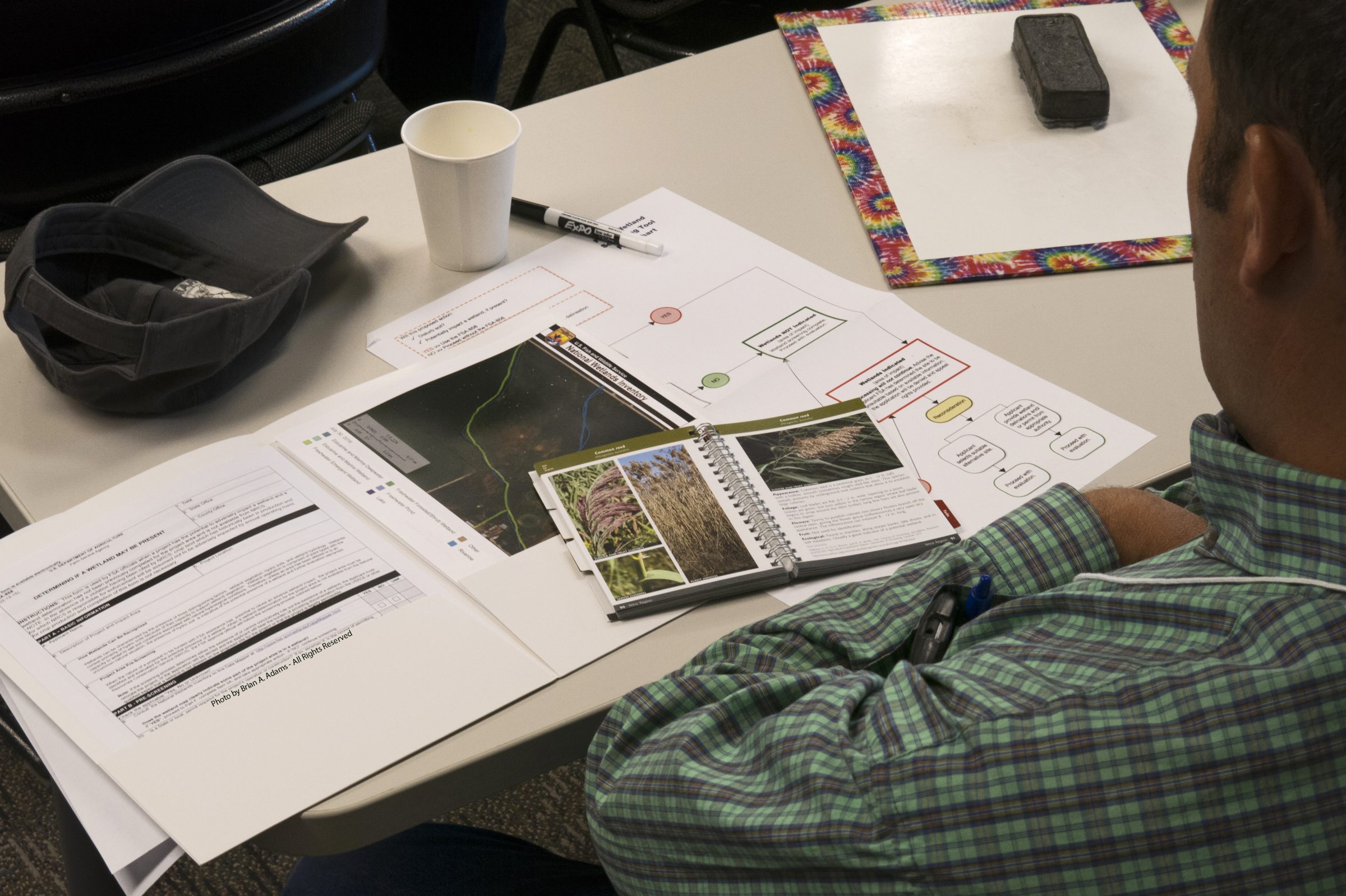 Looking over a persons shoulder to see a coffee of coffee, a hap, and a series of paper materials for a course