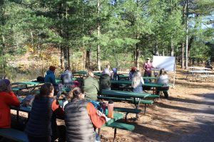 People talking on picnic tables outside in the woods. A person is in front pointing to a white board