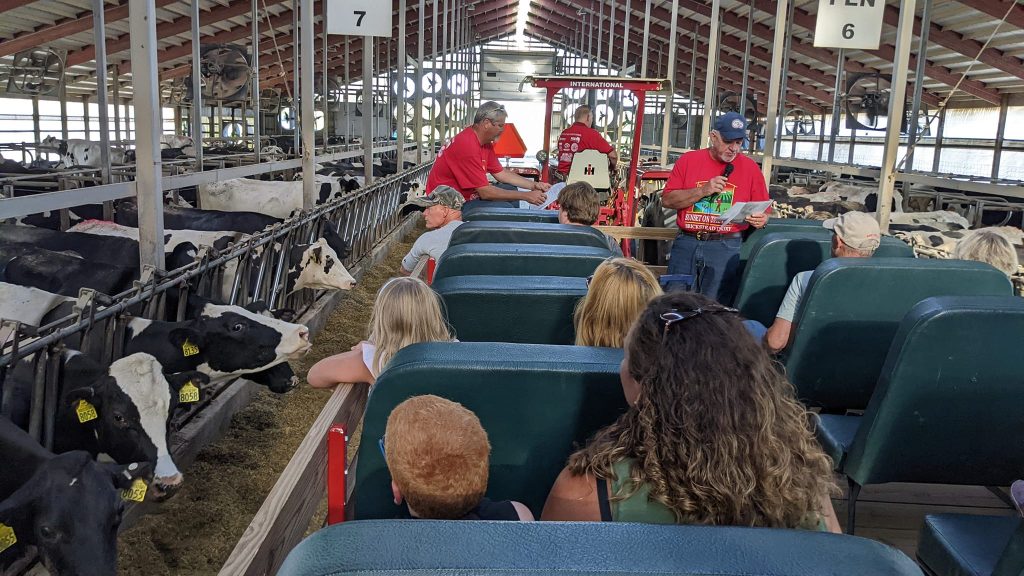 People sitting in a wagon with benches in a dairy barn. A farmer with a microphone is standing and speaking on the wagon.