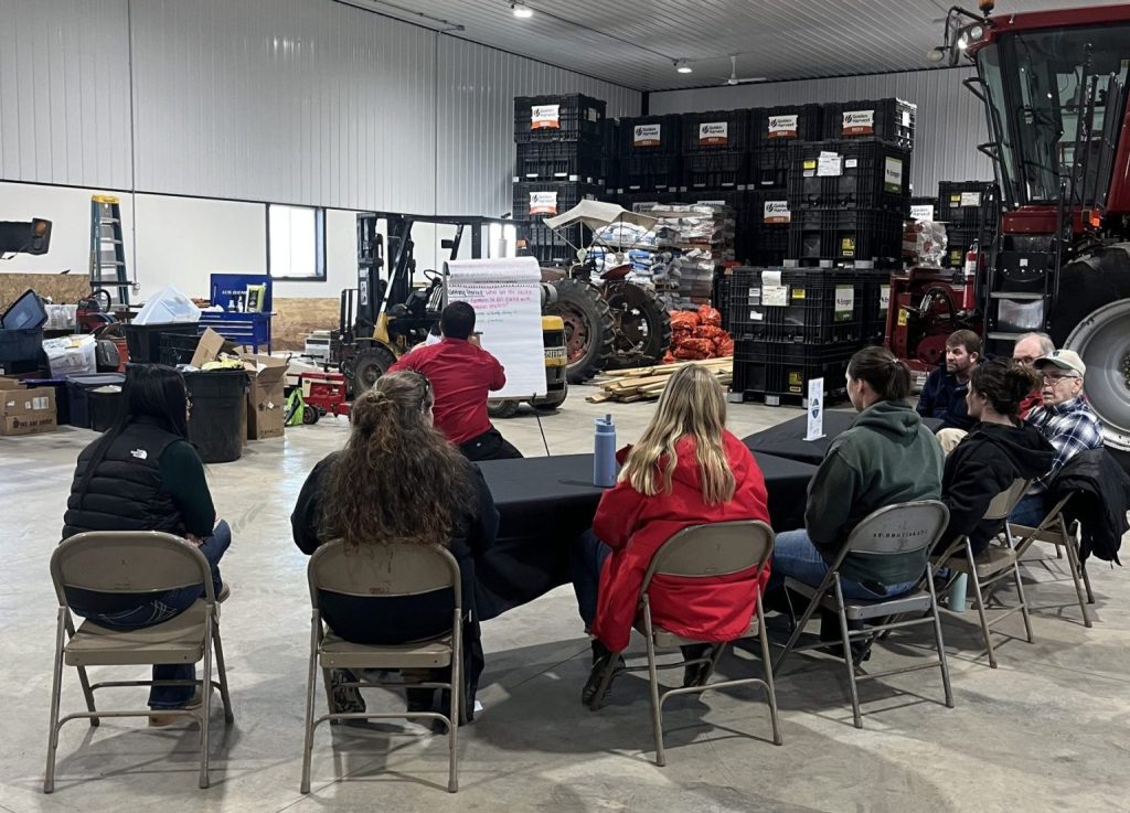 people sitting in a barn on folding chairs listening to a presentation